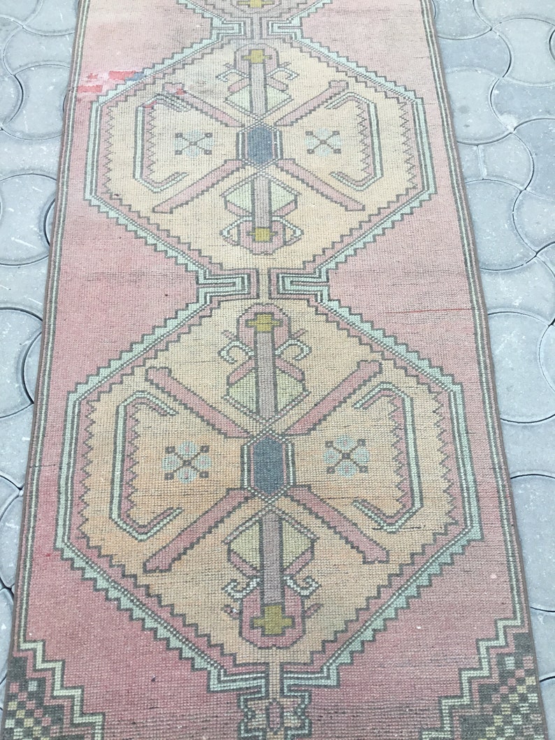 Peut inclure: Un tapis rectangulaire de style vintage, aux tons rose et beige. Il pr&eacute;sente un motif g&eacute;om&eacute;trique central avec des formes r&eacute;p&eacute;t&eacute;es et une bordure en zigzag. Le tapis est probablement en mati&egrave;re tiss&eacute;e.