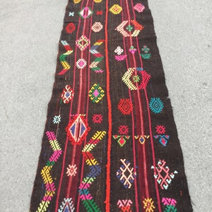 Puede incluir: Una alfombra larga y estrecha con fondo negro y patrones geom&eacute;tricos coloridos. La alfombra tiene una franja roja que recorre el centro y est&aacute; tejida con una variedad de colores, incluyendo rosa, azul, verde, amarillo y blanco.