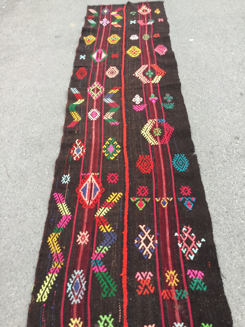 Puede incluir: Una alfombra larga y estrecha con fondo negro y patrones geom&eacute;tricos coloridos. La alfombra tiene una franja roja que recorre el centro y est&aacute; tejida con una variedad de colores, incluyendo rosa, azul, verde, amarillo y blanco.