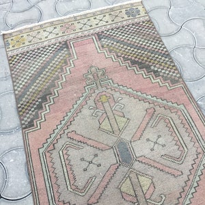 Peut inclure: Un tapis rectangulaire de style vintage avec un motif g&eacute;om&eacute;trique central dans les tons de rose, gris et jaune. Le tapis pr&eacute;sente une bordure d&eacute;taill&eacute;e et des franges sur les bords. Le tapis est pos&eacute; sur une surface grise &agrave; motifs.