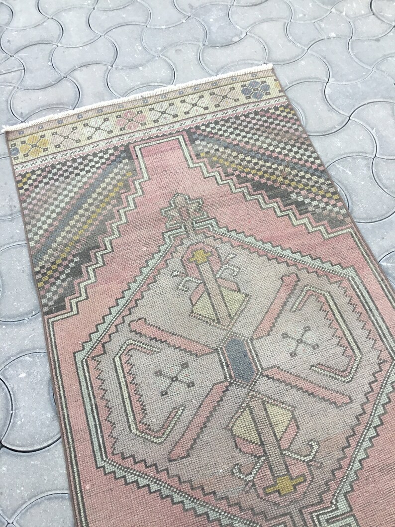Peut inclure: Un tapis rectangulaire de style vintage avec un motif g&eacute;om&eacute;trique central dans les tons de rose, gris et jaune. Le tapis pr&eacute;sente une bordure d&eacute;taill&eacute;e et des franges sur les bords. Le tapis est pos&eacute; sur une surface grise &agrave; motifs.