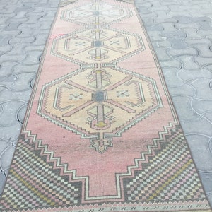 Peut inclure: Un long tapis rectangulaire rose avec un motif g&eacute;om&eacute;trique. Le tapis pr&eacute;sente des motifs de losanges et de zigzags r&eacute;p&eacute;t&eacute;s dans des tons de rose, beige, gris et noir. Le tapis est probablement en laine ou dans une mati&egrave;re similaire.