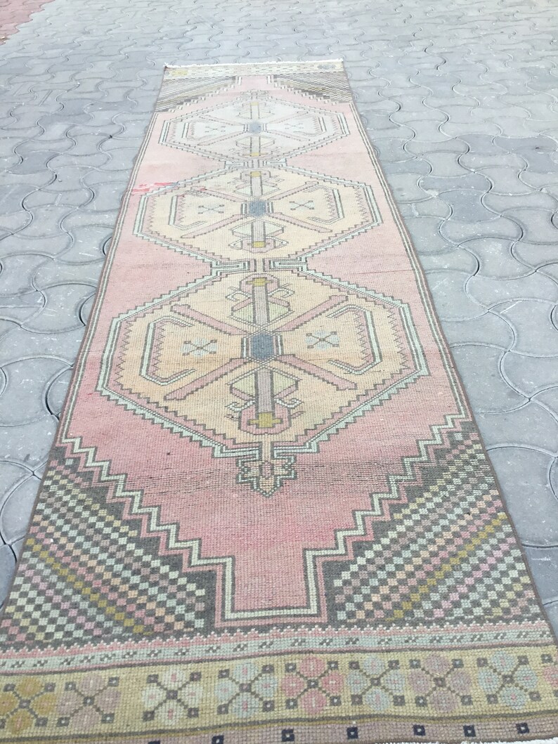 Peut inclure: Un long tapis rectangulaire rose avec un motif g&eacute;om&eacute;trique. Le tapis pr&eacute;sente des motifs de losanges et de zigzags r&eacute;p&eacute;t&eacute;s dans des tons de rose, beige, gris et noir. Le tapis est probablement en laine ou dans une mati&egrave;re similaire.
