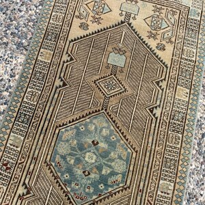 May include: A rectangular woven rug featuring a central blue hexagonal medallion surrounded by intricate geometric patterns in shades of beige, brown, and light blue. The border displays a repeating diamond motif with fringed edges on the ends.