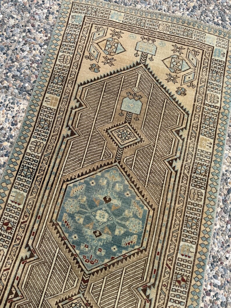 May include: A rectangular woven rug featuring a central blue hexagonal medallion surrounded by intricate geometric patterns in shades of beige, brown, and light blue. The border displays a repeating diamond motif with fringed edges on the ends.