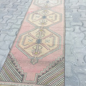Peut inclure: Un long tapis rectangulaire vintage avec un motif g&eacute;om&eacute;trique rose et beige. Le tapis pr&eacute;sente un motif central de formes imbriqu&eacute;es et de bordures avec un m&eacute;lange de couleurs, dont le rose, le beige, le gris et le noir. Le tapis est pos&eacute; sur une surface grise textur&eacute;e.