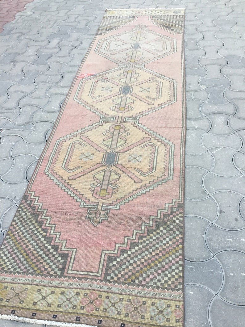 Peut inclure: Un long tapis rectangulaire vintage avec un motif g&eacute;om&eacute;trique rose et beige. Le tapis pr&eacute;sente un motif central de formes imbriqu&eacute;es et de bordures avec un m&eacute;lange de couleurs, dont le rose, le beige, le gris et le noir. Le tapis est pos&eacute; sur une surface grise textur&eacute;e.