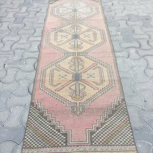 Peut inclure: Un tapis de couloir rectangulaire vintage avec un motif g&eacute;om&eacute;trique rose et beige. Le tapis pr&eacute;sente des formes r&eacute;p&eacute;titives en losange et en croix, avec des accents de noir, de gris et de jaune. Le tapis est pos&eacute; sur une surface grise.