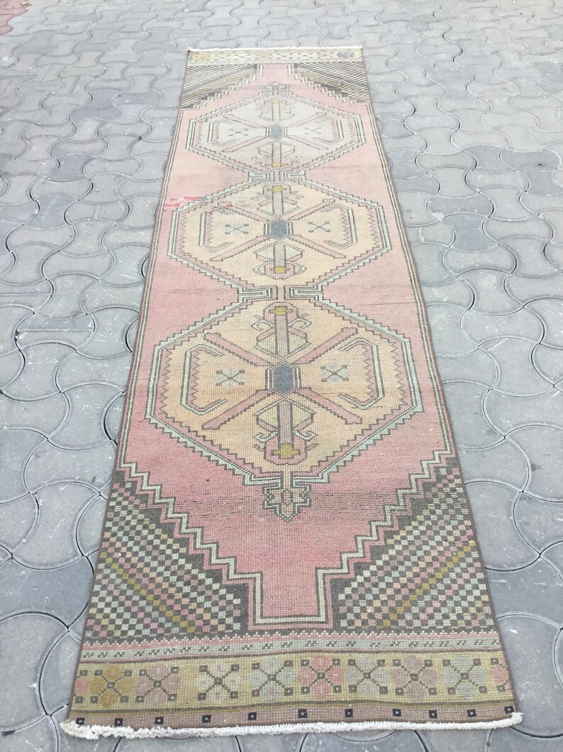 Peut inclure: Un tapis de couloir rectangulaire vintage avec un motif g&eacute;om&eacute;trique rose et beige. Le tapis pr&eacute;sente des formes r&eacute;p&eacute;titives en losange et en croix, avec des accents de noir, de gris et de jaune. Le tapis est pos&eacute; sur une surface grise.