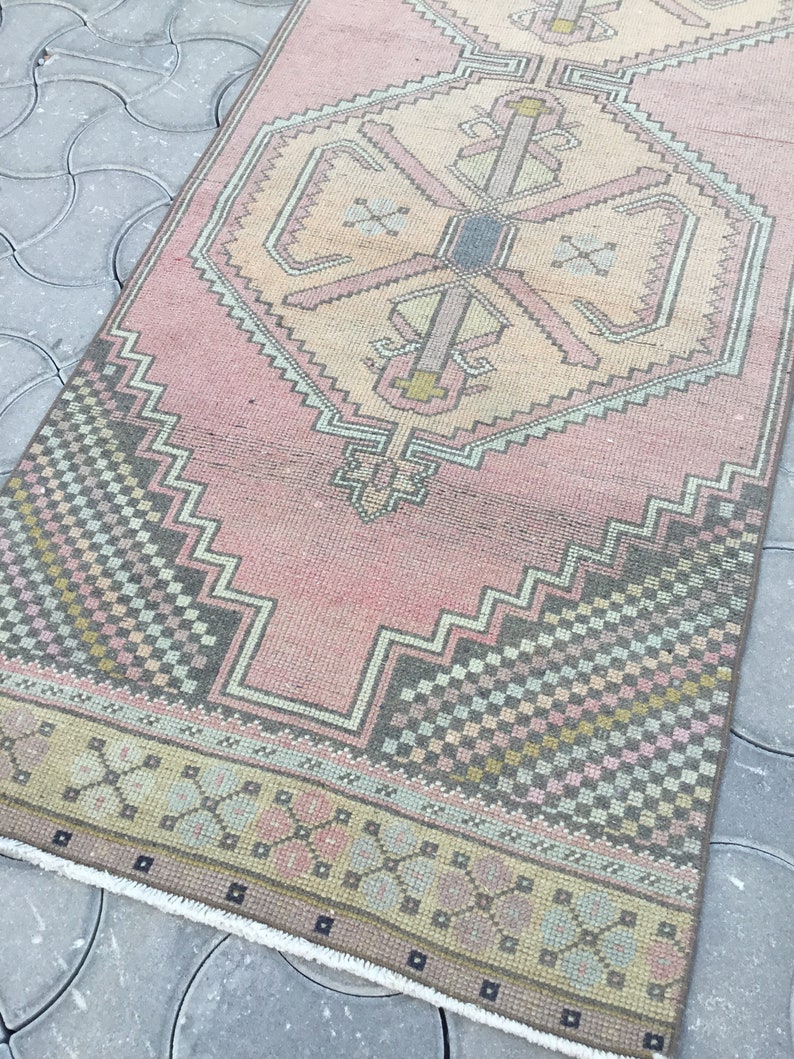 Peut inclure: Un tapis rectangulaire vintage avec un champ central rose et des motifs g&eacute;om&eacute;triques complexes en beige, gris et vert. Le tapis pr&eacute;sente un motif de m&eacute;daillon central et une bordure &agrave; motifs. Les dimensions du tapis ne sont pas visibles.