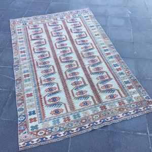 Puede incluir: Una alfombra turca vintage con fondo crema y un patrón geométrico en tonos rojos, azules y marrones. La alfombra tiene un borde con flecos.