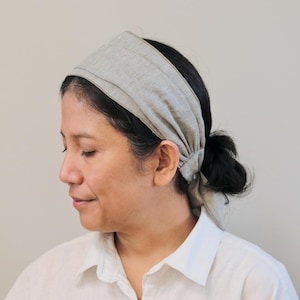 May include: A woman with dark hair is wearing a light gray linen headband. The headband is tied in a knot at the back of her head. She is wearing a white button-down shirt.