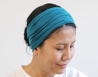 May include: A teal-colored wide headband made of soft fabric. The headband is worn across the forehead and covers the top of the head. The fabric is gathered, creating a textured appearance. The person is wearing a white t-shirt.