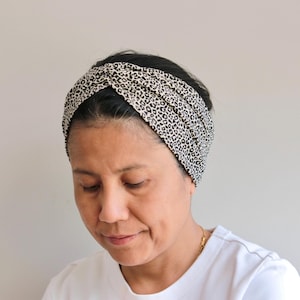 Könnte beinhalten: Ein schwarz-weißes Stirnband mit Leopardenmuster. Der Stoff ist oben am Kopf gewickelt und verdreht. Das Stirnband wird von einer Person mit dunklem Haar und einem weißen Oberteil getragen.