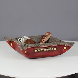 May include: A red leather tray with a gray felt lining. The tray has gold-colored metal accents and the name "MWITOLOLA" is embossed on the front.