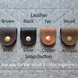 May include: Four leather cable organizers in brown, black, tan, and wood colors, each featuring a metal snap-button in brass, silver, copper, or antique finishes. Text labels identify the leather colors and metal snap-button finishes.