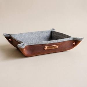 May include: A square valet tray featuring a brown leather exterior and a grey felt interior. The tray is secured at the corners with metallic rivets and includes a small leather tag on the front side with the name FOSTER embossed in capital letters.