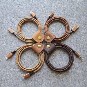 May include: Four coiled charging cables in varying shades of brown, each secured with a leather strap featuring a gold-toned metal snap button. The cables have braided fabric exteriors and metallic connectors at both ends, arranged in a square.