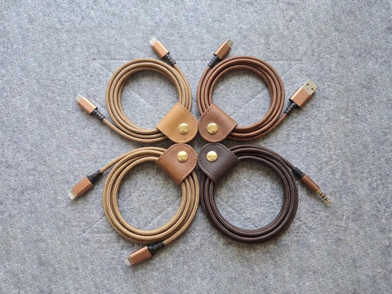 May include: Four coiled charging cables in varying shades of brown, each secured with a leather strap featuring a gold-toned metal snap button. The cables have braided fabric exteriors and metallic connectors at both ends, arranged in a square.