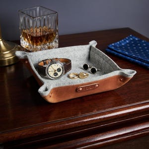 May include: A square valet tray featuring a light gray felt interior and a tan leather exterior with copper-toned metal rivets at the corners. The leather side is embossed with the letters JRL. Inside the tray are a wristwatch, two rings, and cufflinks.