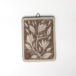 May include: A white ceramic wall hanging with a brown floral design. The design features stylized leaves and flowers.