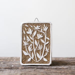 May include: A square ceramic wall hanging with a brown background and a white floral design. The design features stylised leaves and flowers.
