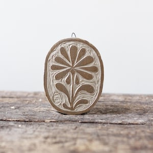 Könnte beinhalten: Ein ovaler Holzblock mit einem geschnitzten Blumenmuster. Das Design zeigt eine stilisierte Blume mit einem zentralen Kreis und Blütenblättern, die sich nach außen erstrecken. Der Block ist hellbraun und hat oben eine Drahtschlaufe zum Aufhängen.