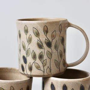 Può includere: Un set di tre tazze in ceramica beige con un motivo a foglie. Le tazze hanno una forma cilindrica con un manico e sono decorate con un motivo di foglie verde oliva e blu navy. Le tazze sono impilate l'una sull'altra.