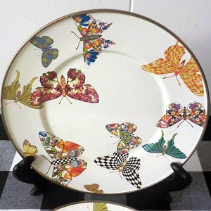 May include: A white ceramic plate with a gold rim, adorned with vibrant butterfly designs. The butterflies showcase diverse patterns and colours, including floral, plaid, and chequered motifs. The plate is presented on a black stand.