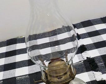 May include: A vintage oil lamp with a clear glass globe and chimney. The lamp has a brass-colored metal base and a round glass reservoir. The lamp is set against a black and white checkered tablecloth.