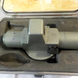 May include: A grey optical level surveying instrument with a cylindrical body and a central housing, featuring a black adjustment knob on the side. The device is securely positioned within a protective, foam-lined carrying case with a metallic frame.