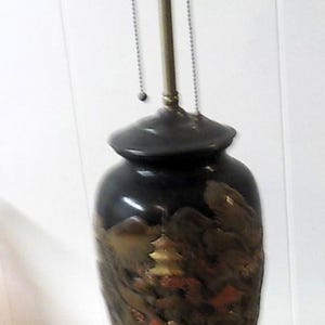 May include: A black table lamp with a decorative vase-shaped base featuring a landscape design in gold, red, and brown. The lamp has three light sockets and a black circular base. The lamp is on a white surface.