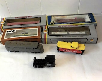 May include: A collection of HO scale model train items including three rectangular cardboard boxes with clear windows, a gray locomotive engine, a yellow and red Rock Island caboose, and a small black steam locomotive engine model.