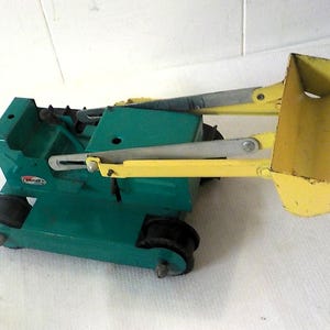 May include: A vintage metal toy tractor with a green body and a yellow front loader bucket. The construction vehicle features black rubber wheels, silver metal support arms for the bucket, and a small red and white logo on the side of the frame.