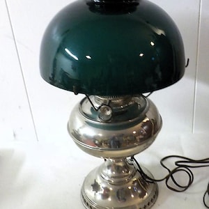 May include: A table lamp featuring a dark green glass dome shade and a polished silver-tone metal base. The lamp has a rounded reservoir, a central burner mechanism with a small adjustment knob, and a perforated decorative metal trim at the bottom.