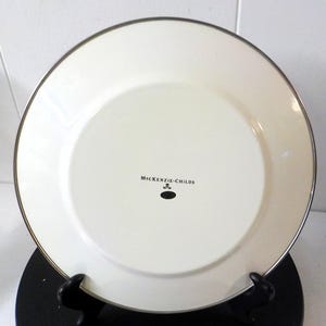 May include: A round, off-white dinner plate with a metallic silver rim. The center of the plate features the black text MacKenzie-Childs printed above a small black oval icon. The plate has a smooth, glossy finish and a simple, clean design.