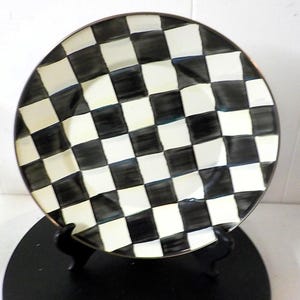 May include: A round ceramic plate featuring a hand-painted black and white checkered pattern. The squares have a slightly irregular, brush-stroke appearance, and the plate has a thin, dark metallic-toned rim around the outer edge.