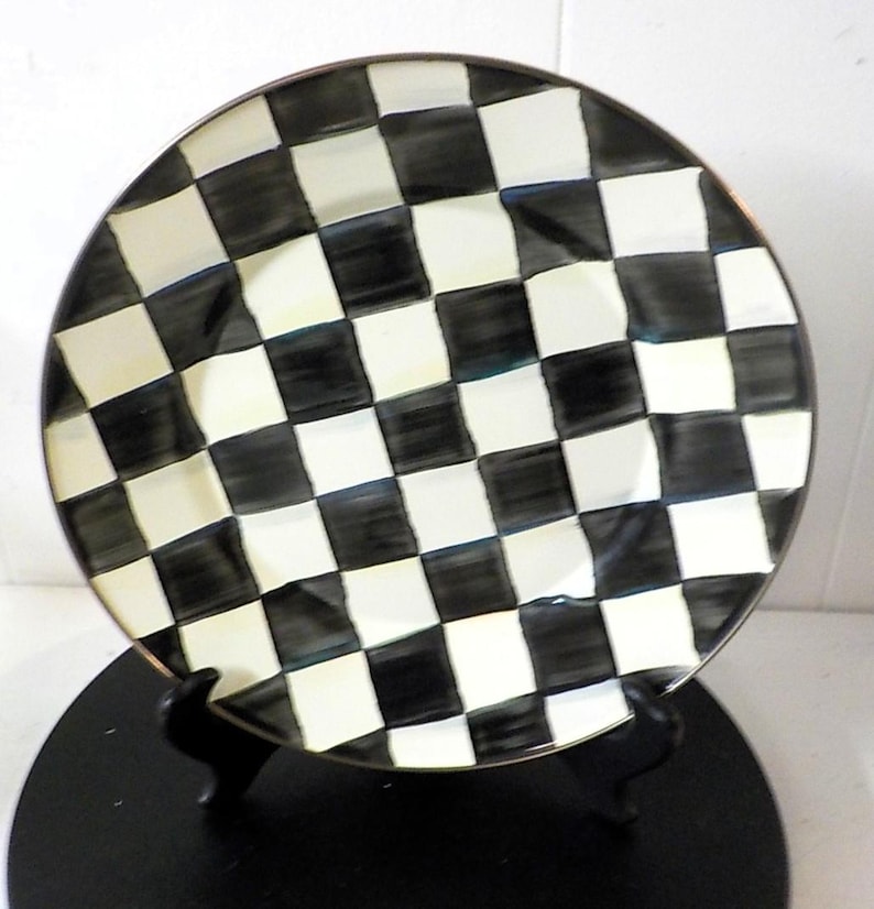 May include: A round ceramic plate featuring a hand-painted black and white checkered pattern. The squares have a slightly irregular, brush-stroke appearance, and the plate has a thin, dark metallic-toned rim around the outer edge.