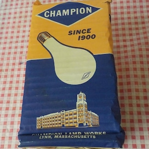 Puede incluir: Caja de bombilla Champion vintage. La caja es principalmente amarilla y azul, con un gran gráfico de bombilla y el texto "CHAMPION SINCE 1900". La parte inferior de la caja muestra una ilustración de una fábrica con el texto "CHAMPION LAMP WORKS LYNN, MASSACHUSETTS."