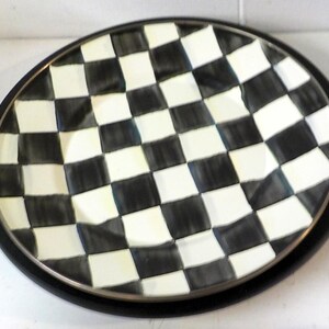 May include: A round ceramic plate featuring a hand-painted black and white checkered pattern across the entire surface. The plate has a slightly wavy, uneven rim and a solid black outer edge, creating a bold contrast between the light and dark squares.