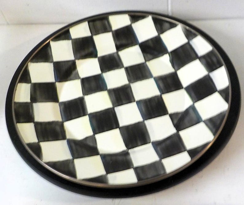 May include: A round ceramic plate featuring a hand-painted black and white checkered pattern across the entire surface. The plate has a slightly wavy, uneven rim and a solid black outer edge, creating a bold contrast between the light and dark squares.