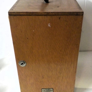 May include: A rectangular wooden box with a black handle on top. The box is made of light brown wood and has a metal latch on the front. The box has the word "OLYMPUS" on a small metal plate.