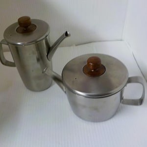 May include: Two stainless steel teapots with wooden knobs on the lids. One teapot is tall and cylindrical, while the other is shorter and rounder. Both have spouts and handles, and are set against a white background.