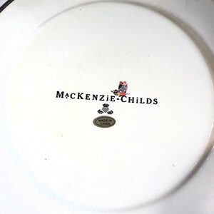 May include: White ceramic plate with a silver rim. The plate features the "MacKenzie-Childs" logo in black, a colourful butterfly design, and a small "Made in China" sticker. The plate is likely for decorative or dining use.