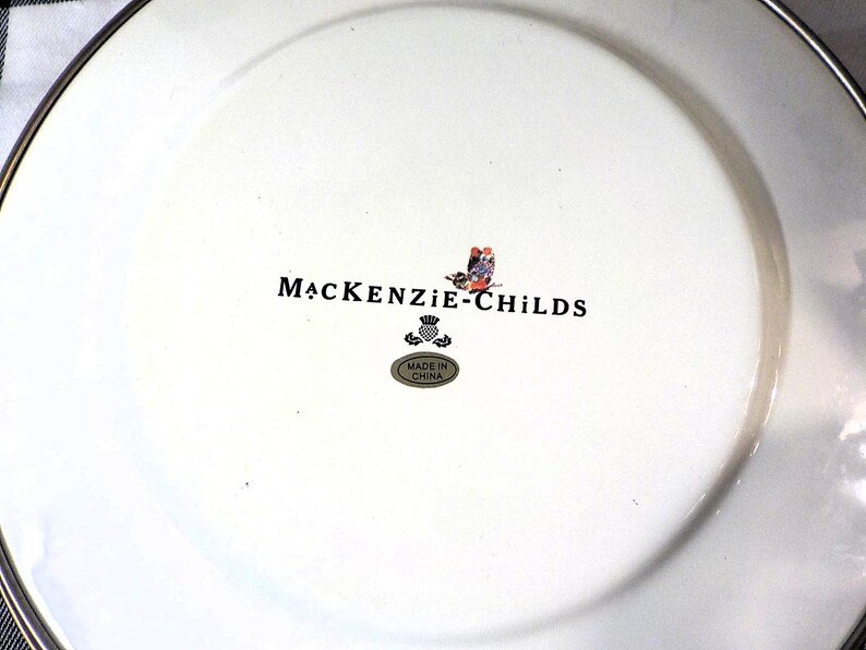 May include: White ceramic plate with a silver rim. The plate features the "MacKenzie-Childs" logo in black, a colourful butterfly design, and a small "Made in China" sticker. The plate is likely for decorative or dining use.