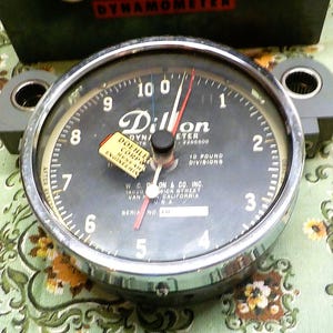 May include: A Dillon dynamometer with a black face, silver bezel, and gray mounting brackets. The gauge reads up to 10, with a red needle and a small yellow sticker. The text "Dillon" and "18 Pound Divisions" are visible.