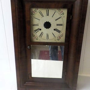 May include: A rectangular wall clock with a dark brown wooden frame. The upper section features a cream-colored face with black Roman numerals and floral accents in the corners. The lower section contains a rectangular mirror panel with a small knob.