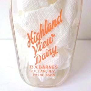 May include: A clear glass Highland View Dairy milk bottle with orange lettering. The bottle contains white paper and has the text "B.V. Barnes, Olean, N.Y., Phone 7666" at the bottom.