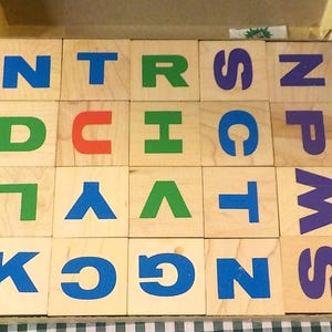 May include: A set of twenty square wooden blocks arranged in a four-by-five grid inside a cardboard box. Each block features a single large letter printed in blue, green, red, or purple ink. The letters include N, T, R, S, P, D, U, H, C, M, L, Y, V, T, S, K, C, G, Z, and S.