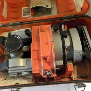 May include: A grey surveying instrument with black adjustment knobs and a circular lens, housed inside a bright orange protective carrying case. The device features metallic components and a central orange frame, secured within the molded interior.
